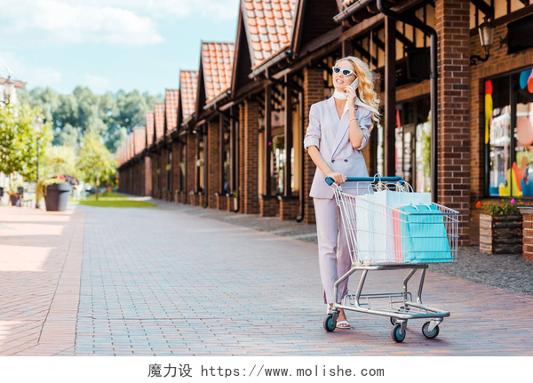 一个女孩推着购物篮打电话美丽的年轻女子与购物车满纸袋聊天电话在街上
